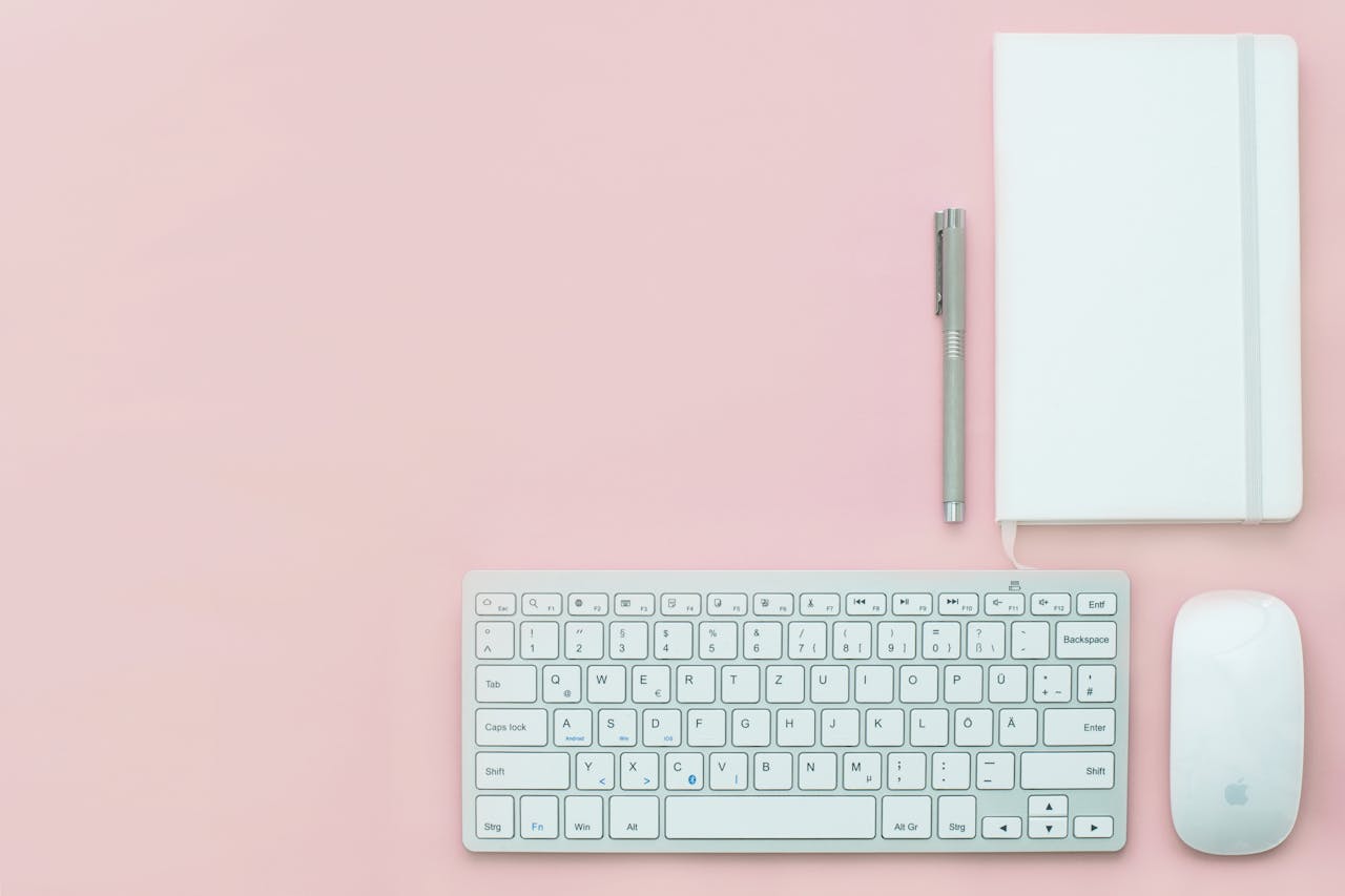Flat lay of a minimalist workspace featuring a keyboard, notebook, and pen on a pink surface.